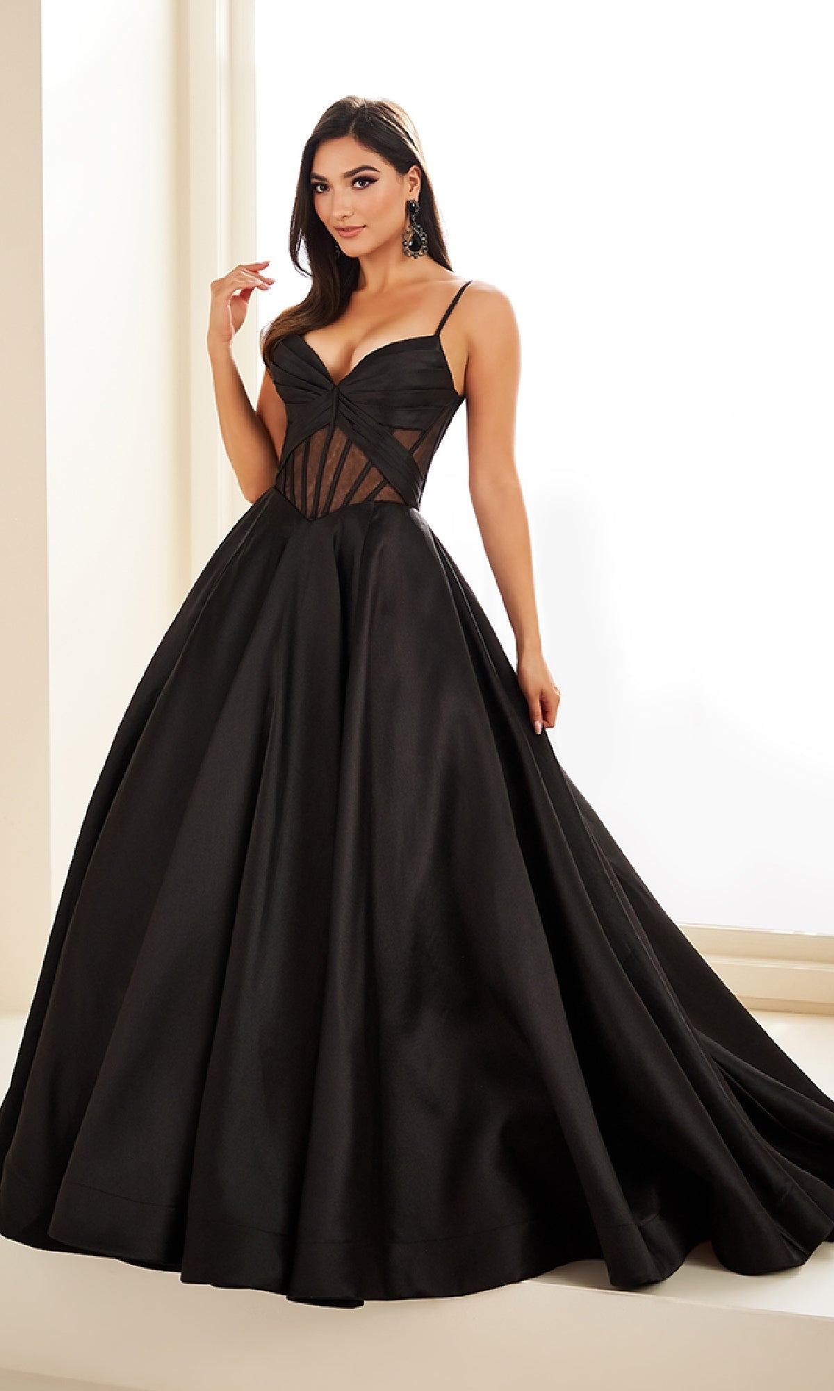 A woman poses indoors in the Sheer-Corset Prom Ball Gown: Ellie Wilde EW36073, a long black dress with pockets, fitted bodice, sheer panels, and sweetheart neckline. She gracefully holds the skirt with one hand.